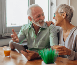 Senior man and woman sitting at table laughing holding a tablet