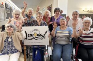 518318939_1296188072514246_3460695217953563520_n - Grace Management Smiling group of senior women, some in wheelchairs, raise their hands behind a banner that says Payton's Lemonade Stand, Kids Serving Kids.