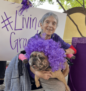 Smiling elderly woman in a wheelchair hugs a bulldog wearing a purple curly wig, beside a banner that reads '#Living' in purple.