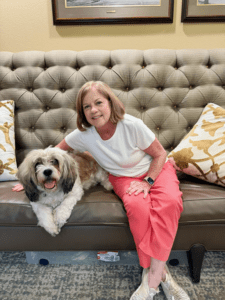 Older woman in a white top and coral pants sits on a tufted beige sofa with a fluffy white and gray dog beside her, in a living room setting.