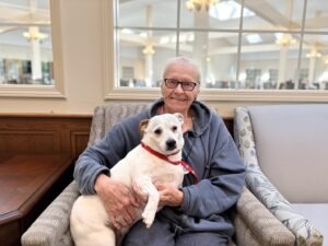 Smiling senior woman with glasses sits in a patterned chair, holding a small white dog with a red collar in a bright lounge setting.