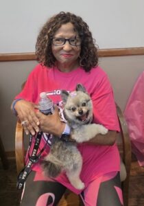 Smiling woman with curly hair and blue glasses wearing a bright pink shirt sits in a chair, holding a small fluffy dog and a water bottle.