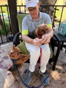 Older man in a polo and cap sits outdoors on a patio, holding a small brown dog while another small dog on a blue leash stands beside him.