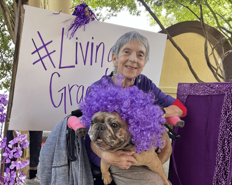 featured image - Grace Management Smiling elderly woman in a wheelchair hugging a bulldog wearing a bright purple curly wig at an outdoor purple-themed event, with a white sign in the background reading '#Living Grace'.