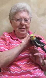 Elderly woman with white hair in a pink chevron shirt holding a yellow- and gray cockatiel on her finger, smiling at the bird.