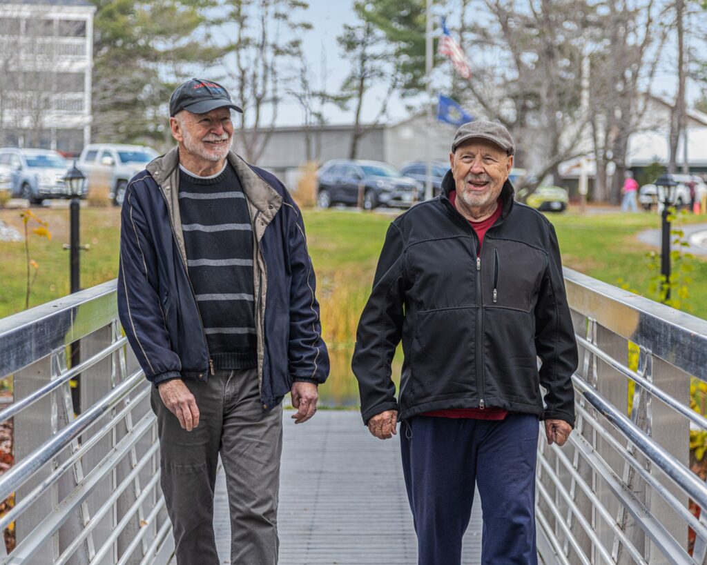 Two elderly men walk side by side on a metal pedestrian bridge, smiling with a park and cars in the background.