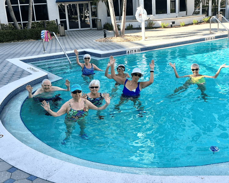 Six seniors in a sunny pool, waving at the camera while standing in the water, wearing swimsuits and hats.